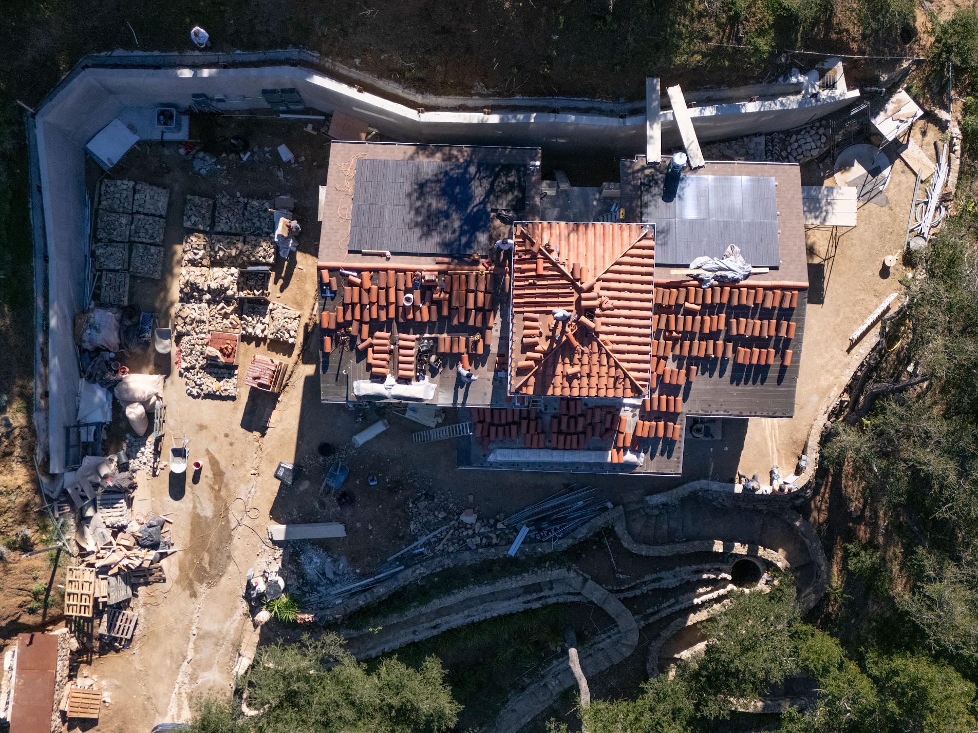 Drone view straight down on the roof, with rows of clay tiles, a partly tiled hip roof, two solar arrays, and stacks of stone and building materials on the ground.