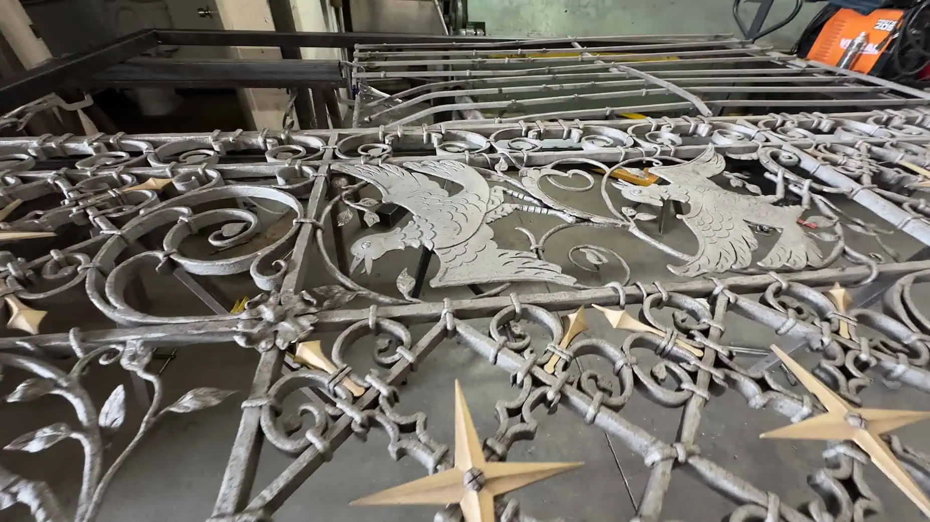 Wrought iron gate panels laid out flat on a shop floor, showing bird figures, scrollwork, and gold star-shaped finials, with steel stock and equipment in the background.
