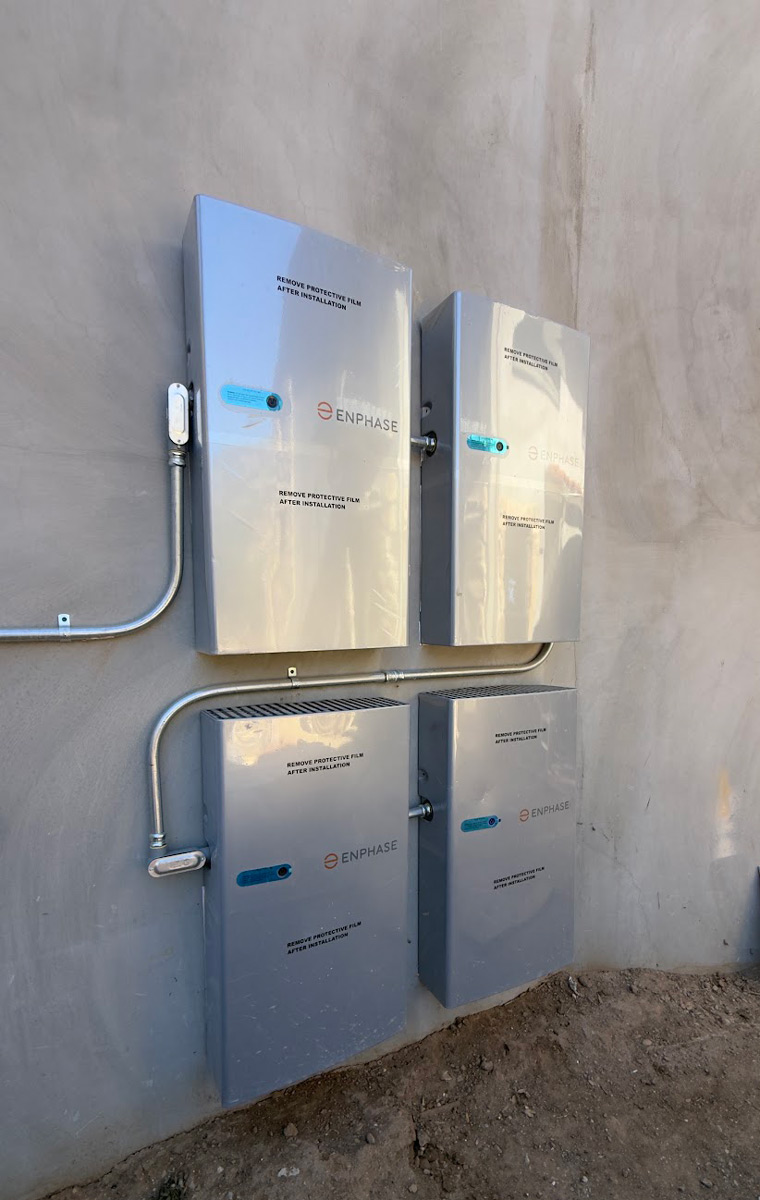 Four silver Enphase battery units mounted in two rows on a stucco wall, still wearing protective film and joined by metal conduit.