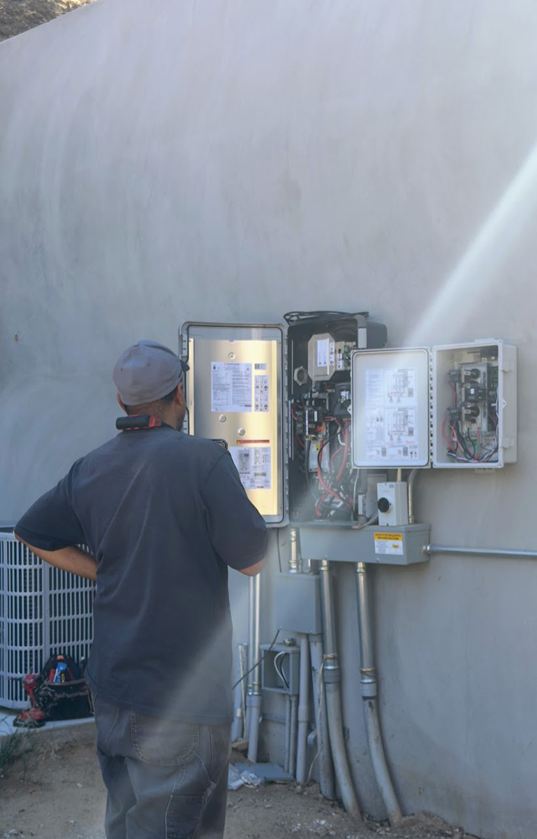A technician in a gray cap, hand on hip, looks at the open Enphase electrical panels on a stucco wall.