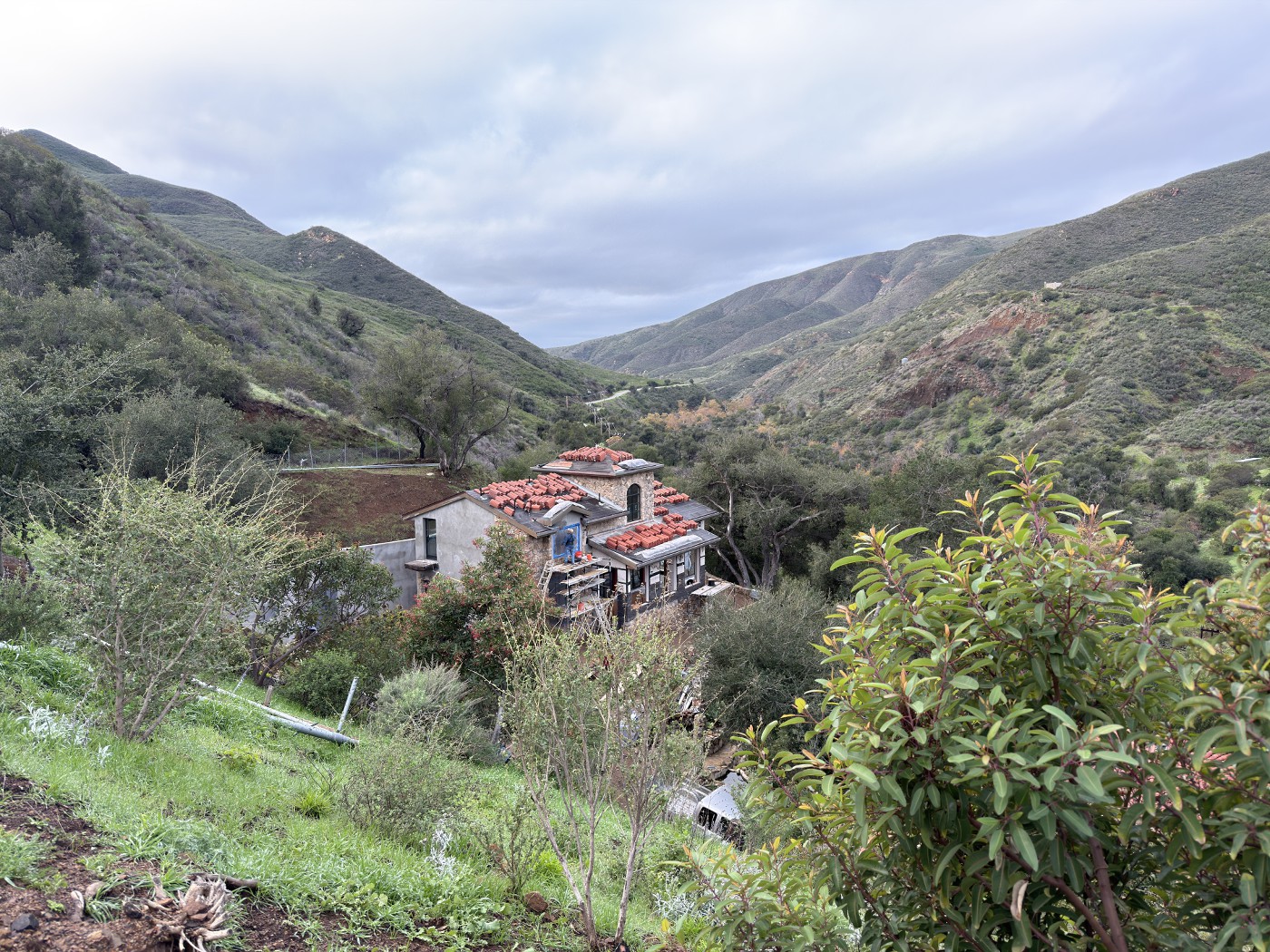 Wide view of green hills under an overcast sky, with the partly tiled house in the valley below.