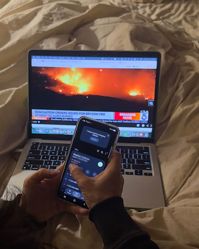 Diane checking her phone in front of a laptop showing a KTLA breaking-news report of Bryson Fire evacuation orders, with flames on the hillside.