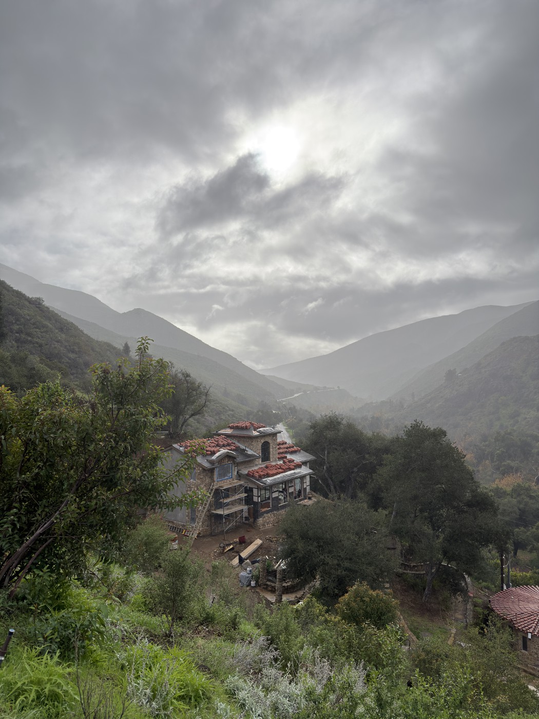 Stone house with scaffolding in a misty canyon under dark clouds, with tile stacks on the roofs.