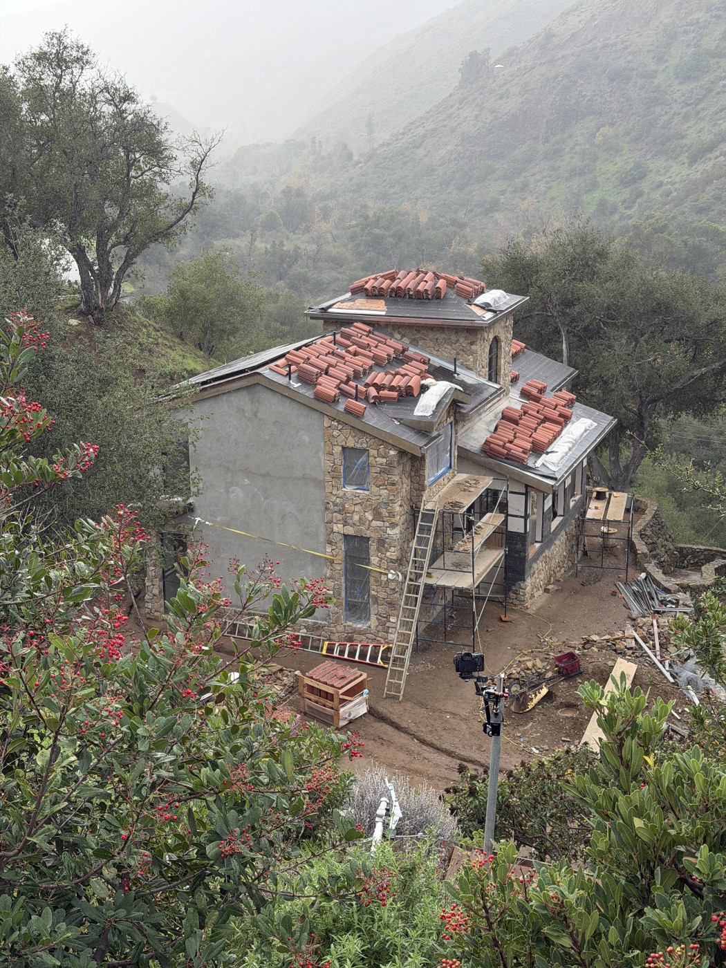 Stone and plaster house in fog, with stacks of clay tiles on dark roof underlayment, a ladder and scaffolding in front, and misty hills behind.