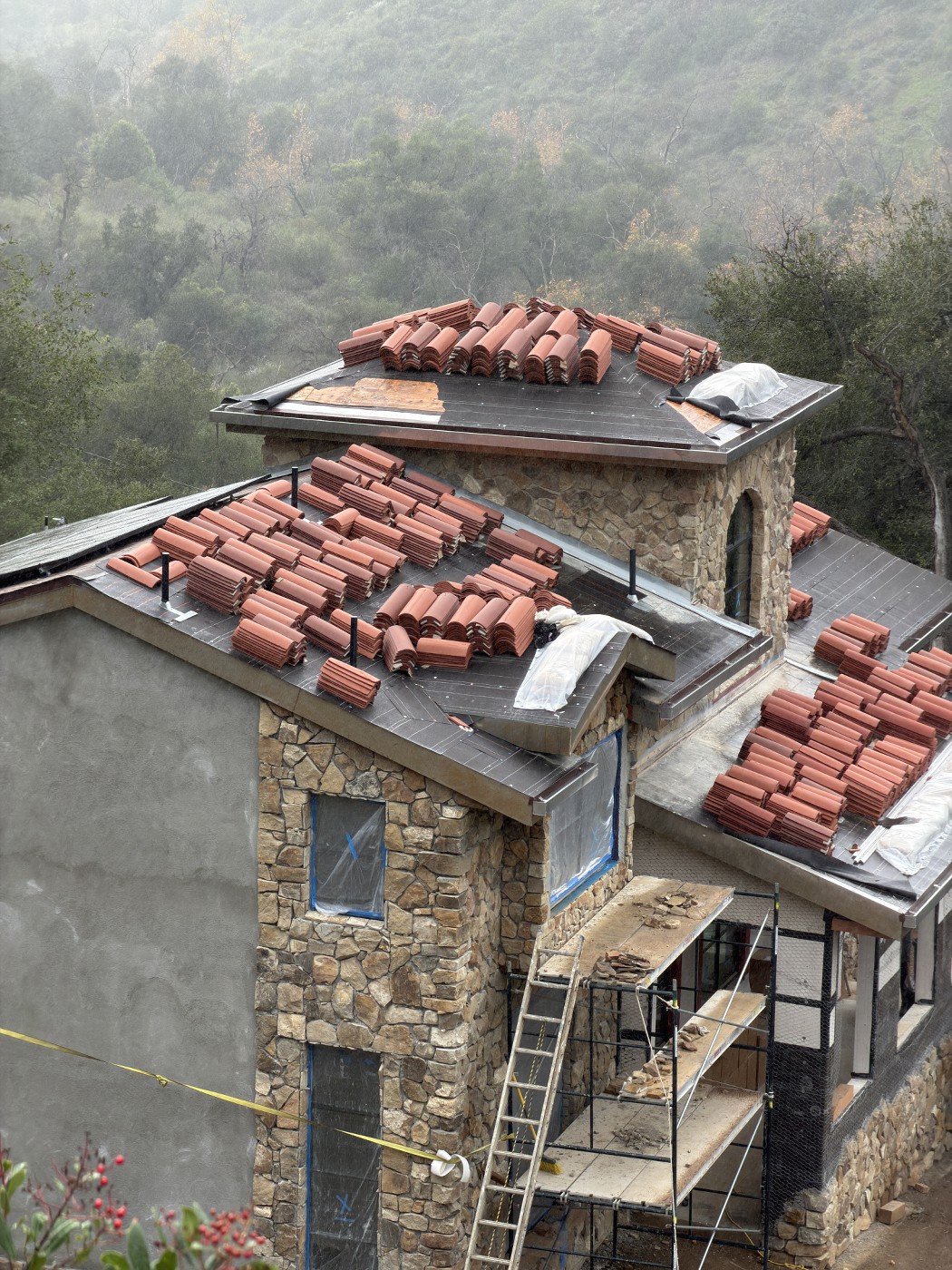 Working during the rain. Close view of stacked red clay tiles on a dark underlayment, next to a stone tower with an arched window.