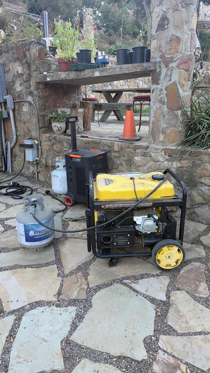 A yellow portable dual fuel generator on wheels and two propane tanks on a flagstone patio, with a Jackery power station and a stone wall behind.