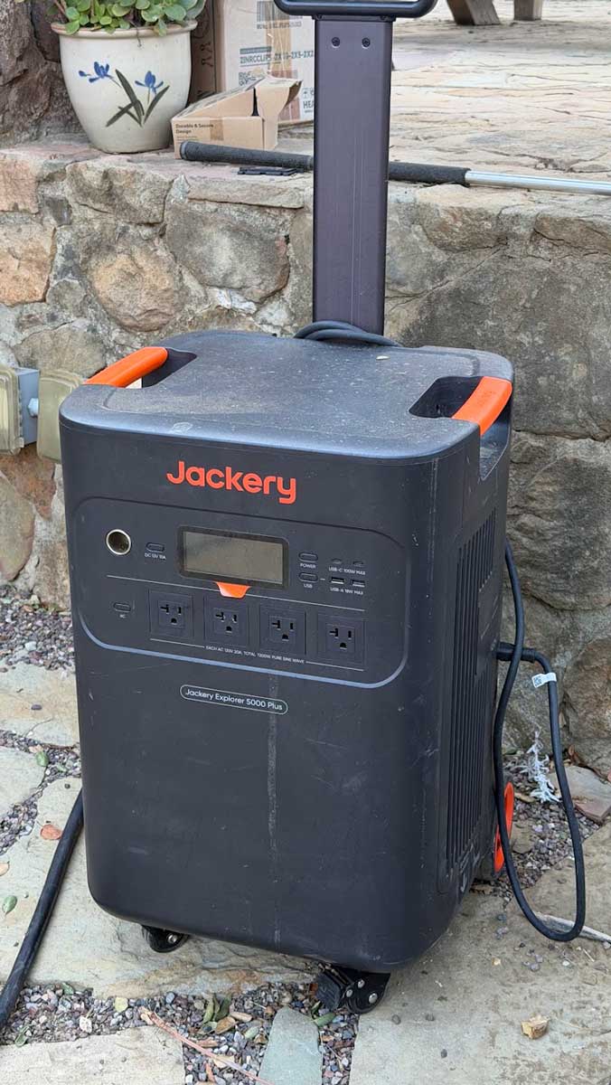 A black Jackery Explorer 5000 Plus portable power station with orange handles, on wheels, beside a stone wall on a flagstone patio.