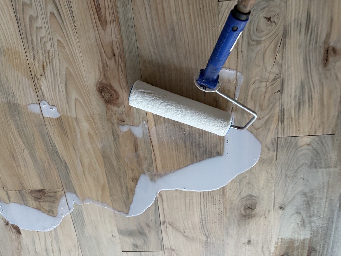 Close view of wood planks with knots, a paint roller lying on the floor beside a spill of milky white finish.
