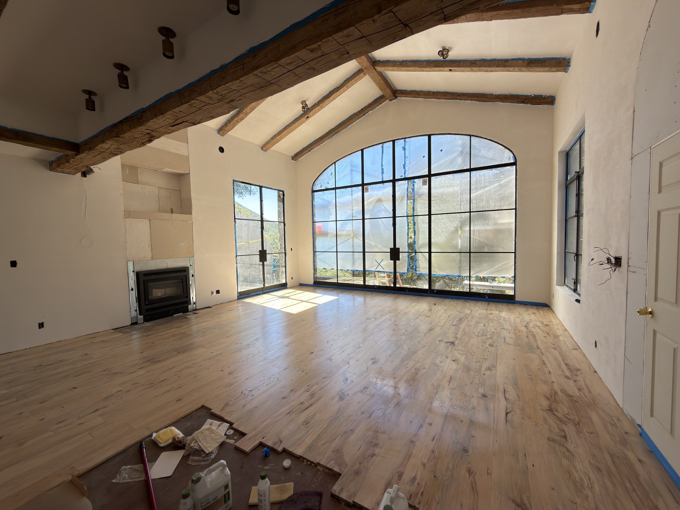 Wood floor with a glossy finish in a room with exposed beams, a fireplace insert and a tall arched window, with finishing supplies on paper in the foreground.