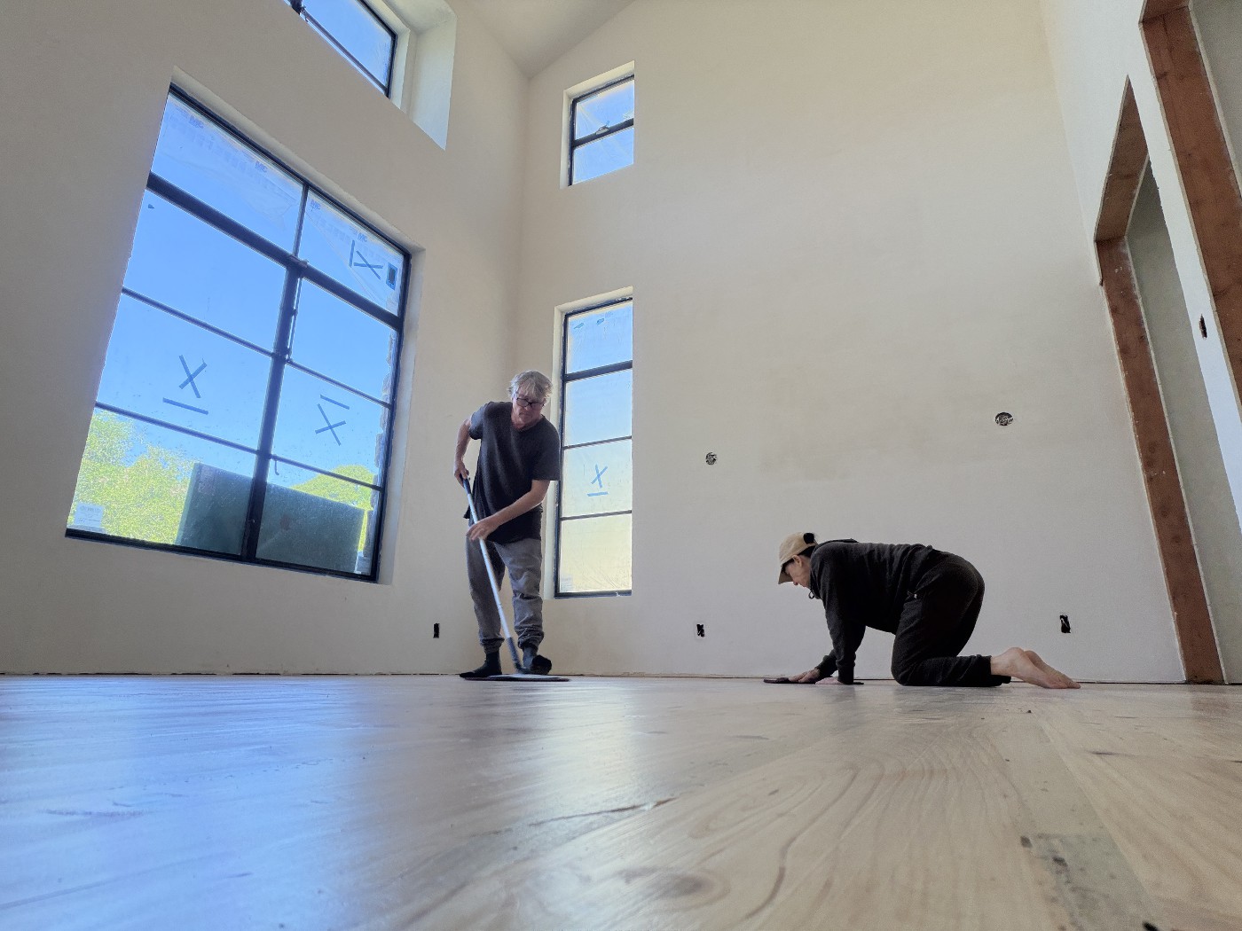 Two people working on a pale wood floor in a tall room with large windows, one standing with a long-handled applicator and one kneeling