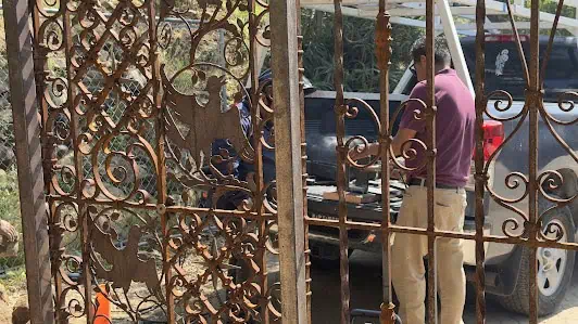 Rust-colored wrought iron gate with scrolls and bird shapes in the foreground, and a man in a maroon shirt working beside a parked vehicle behind it.