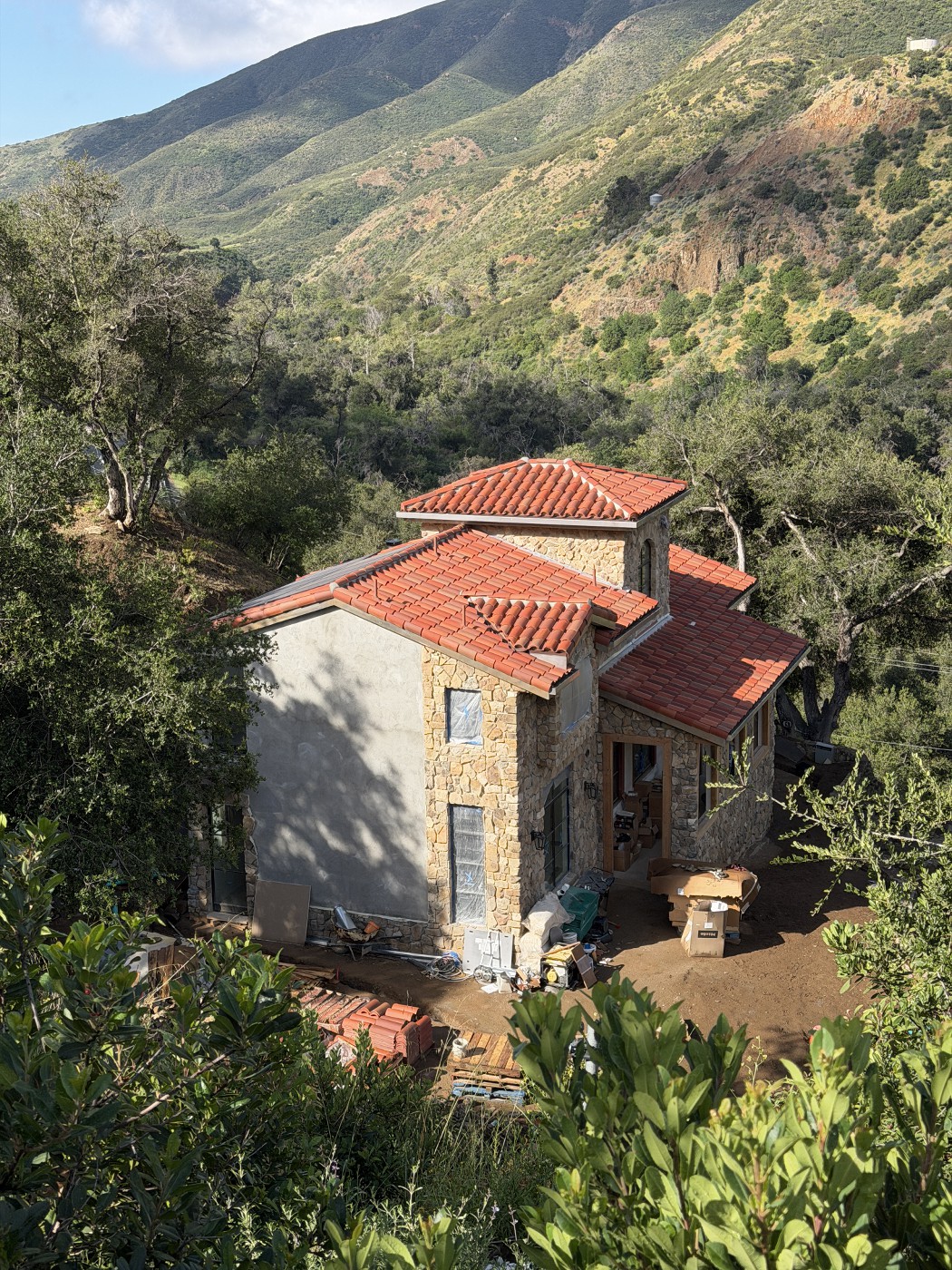 Stone and plaster house with a finished red clay tile roof and tower, surrounded by oak trees and green hills.