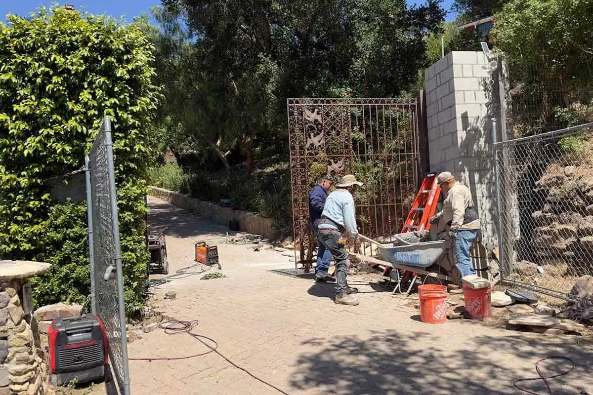 Three workers at the driveway gate, one holding a wheelbarrow of mortar, with flagstone stacked on the ground and a ladder against the cinder block pillar.