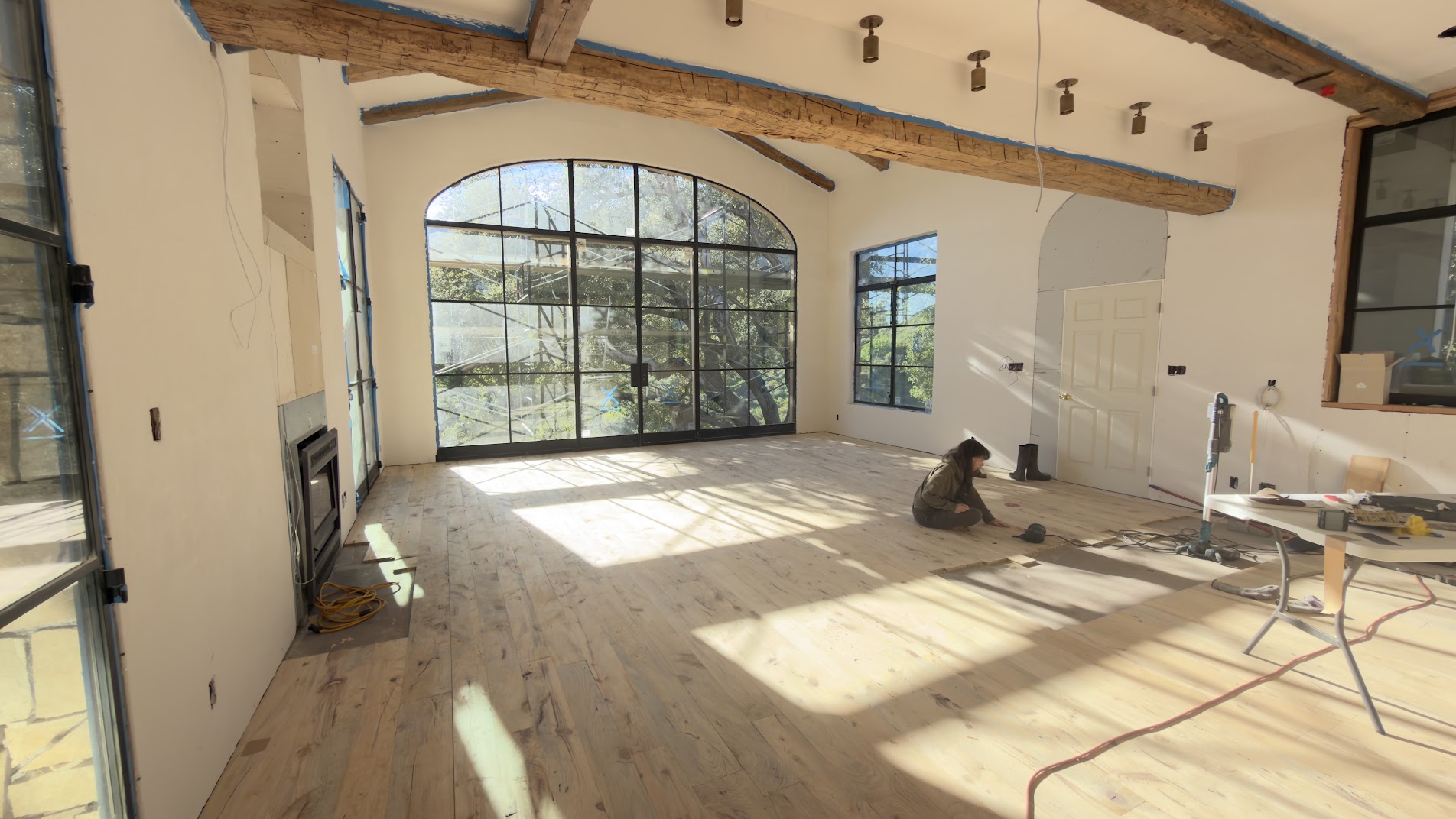 Sunlit room with beams and an arched window, a pale wood floor mostly laid, a bare patch to the right of center, and a person kneeling on the boards.