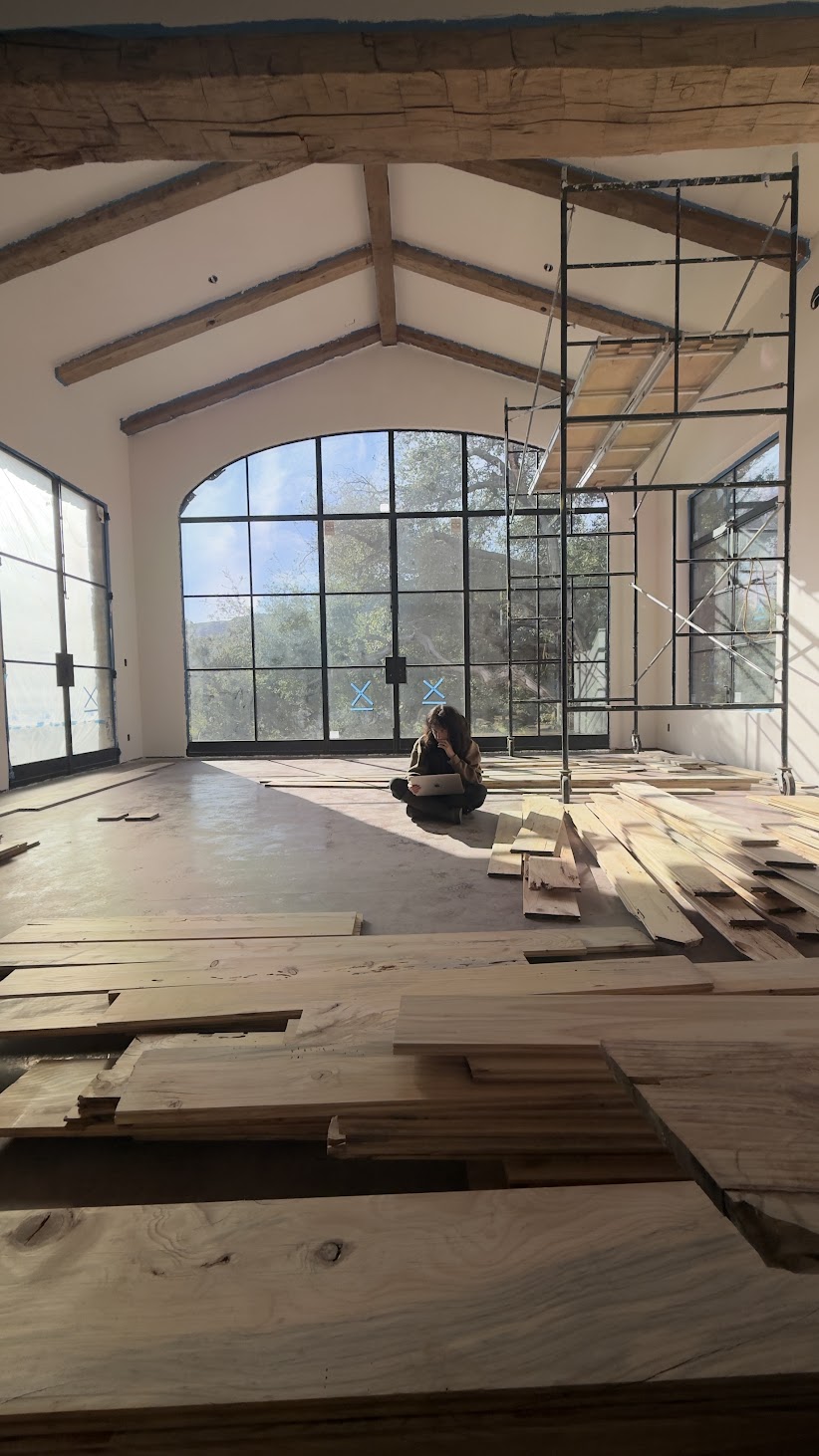 Large room with exposed beams and an arched window, piles of wood boards in the foreground, a person sitting on the floor with a laptop, and a scaffold tower at right.