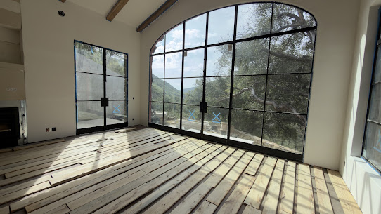 Long wood boards laid loosely in rows on a bare floor in front of a tall arched steel-framed window with oak trees outside.