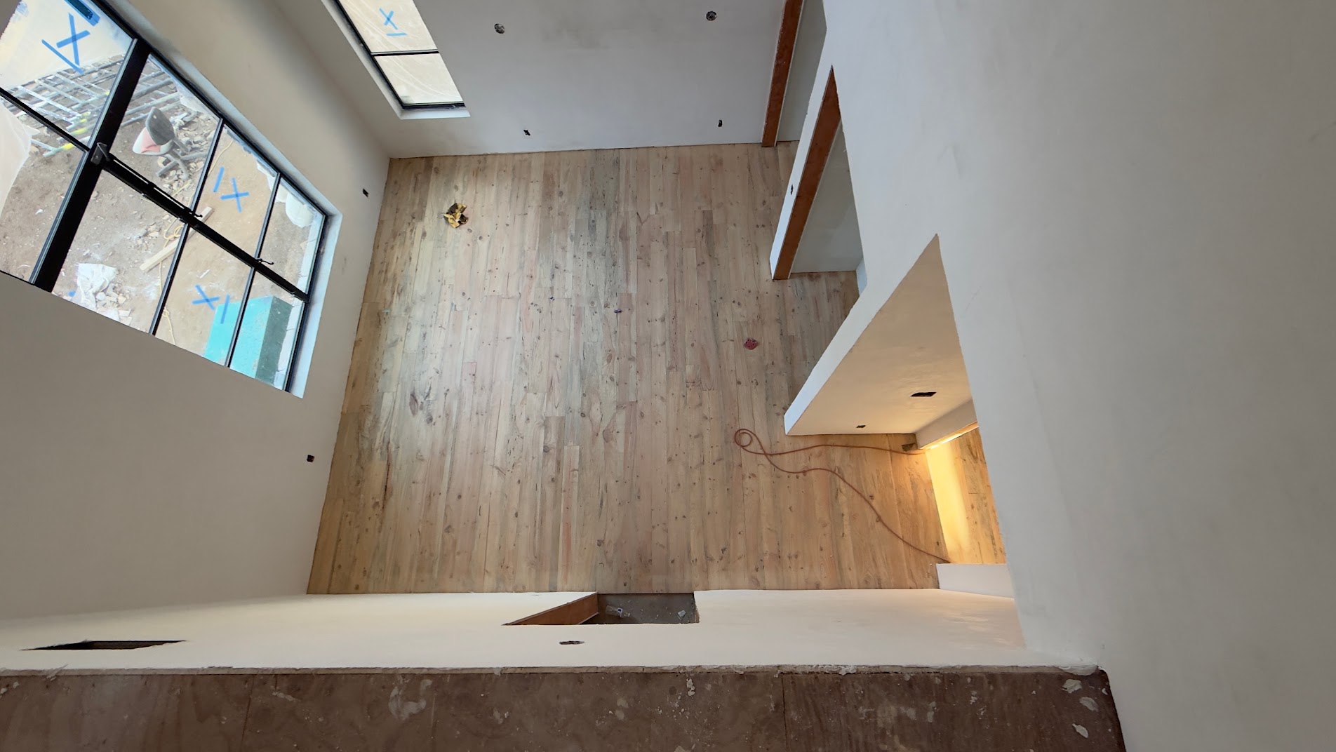 View down from a loft onto a sanded wood floor in a room with a large window, an extension cord lying across the boards.